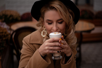 Beautiful european woman sit in the terrace of cafe in france style , holding a cup of latte with airy milk froth . The female enjoy the retro dtreet of the city. Model wearing coat and hat