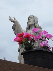 Scene in a cemetery: in the foreground, a black plastic vase with artificial pink flowers. Behind, unfocused, a statue of Jesus Christ with his arm raised and looking up at the cloudy sky. 