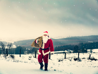 Santa Claus with a bag full of Christmas presents is walking along a snowy field to people for a...