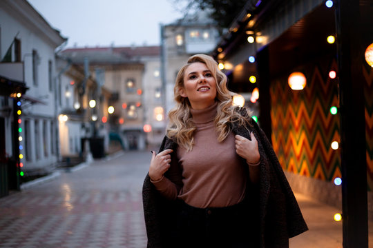 Fashion woman walking at the city, dressed modern clothes. Female model wearing coat, tutleneck, trousers and posing against the street with light at the evening