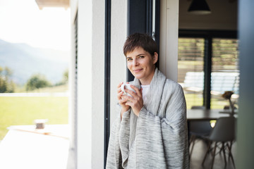 A young woman with coffee standing by patio door at home.