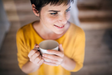 Front view of young woman with coffee standing indoors at home.