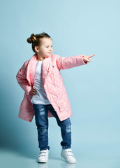 Stylish kid girl in long winter jacket white t-shirt, modern blue torn jeans is pointing her finger aside