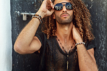 Handsome man with curly hair and jewelry on his arms and chest, outdoor photoshoot under the sun - image
