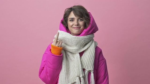 Attractive young woman wearing a winter jacket with a scarf is showing finger middle isolated over pink background