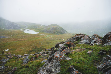Beautiful snowfall in mountains. Atmospheric green alpine landscape with large snowflakes. Small lake, water streams and rocky hill with sharp craggy stones during snowfall. Flakes of snow in highland