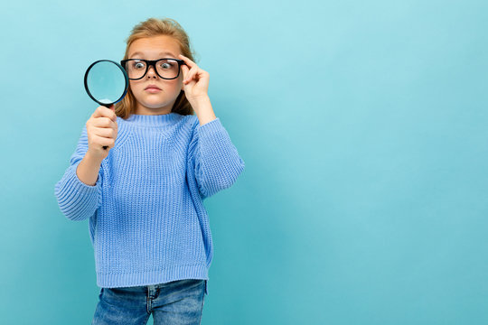 surprised european girl in glasses looks through a magnifying glass on a light blue background with copyspace