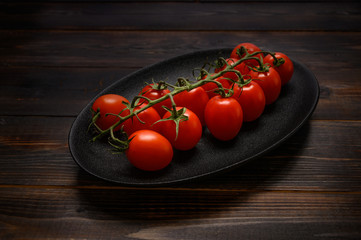 Red tomatoes on a green branch on a dark platter on a wooden background. Rustic style. Top view