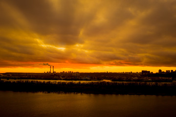 Epic sunbeams through thunderclouds in the bright sky during sunset over the city river. Sunset clouds in a sunset city over a river.