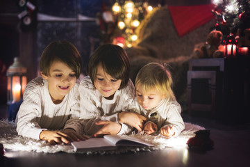 Three children, boy brothers, reading book at home at night on Christmas evening
