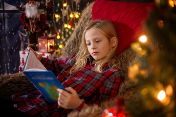 Cute preschool girl sitting around christmas tree in cozy chair, reading