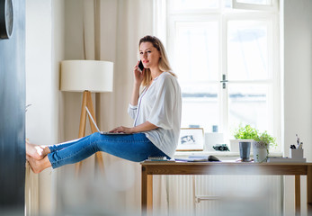Young woman with smartphone and laptop indoors in home office, working.