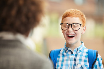 Happy schoolboy in eyeglasses laughing while standing and talking to his friend