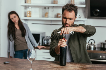 Selective focus of alcohol depended man opening wine bottle and worried wife on kitchen