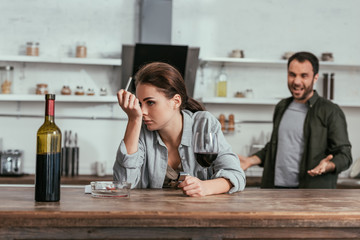 Selective focus of woman with cigarette and wine and quarreling man on kitchen