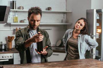 Selective focus of alcohol depended man with wine bottle and sad wife on kitchen