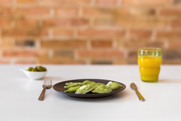 Traditional russian and ukrainian vegetarian green vareniki dumplings on a white plate, with olives and orange juice. Dough colored with spinach juice. Brick wall as the background.