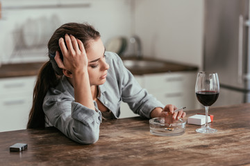 Worried woman smoking cigarette beside wine glass on table