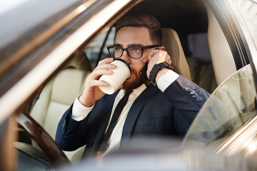Young businessman in eyeglasses drinking coffee and talking on mobile phone in the car