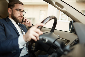 Young serious businessman in eyeglasses talking on mobile phone and driving the car