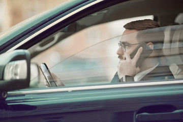 Side view of young businessman in eyeglasses talking on mobile phone and watching for the road in the car