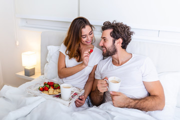 Cute couple having breakfast in bed in the bedroom. Beautiful woman feeding her boyfriend strawberries in bed while having breakfast and coffee in bedroom - obrazy, fototapety, plakaty