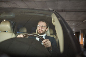 Young serious man in eyeglasses holding the wheel and sitting in his car in the parking
