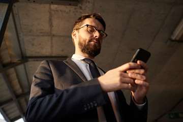 Low angle view of bearded businessman in suit standing and working online using his mobile phone