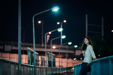 asian young woman with long hair in white top and jeans standing alone on the bridge at night with eyes on the camera feeling lonely and missing someone