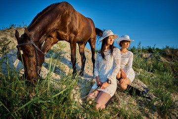 two women in white dresses and white hats on a summer evening with a horse at sunset