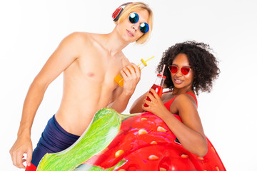 Pretty african female and caucasian blonde man stands in swimsuit with rubber beach mattresses, drinks juice and listen to music isolated on white background