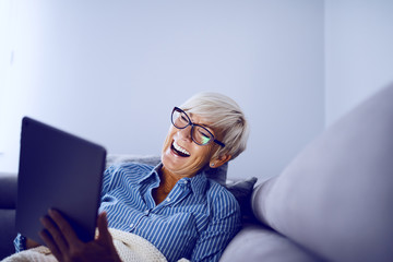 Attractive caucasian smiling blonde senior woman sitting on sofa in living room, using tablet and...