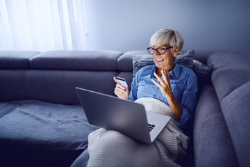 Cheerful charming caucasian senior blond woman with short hair and with eyeglasses sitting on sofa in living room, holding laptop in lap and using credit card for online shopping.