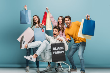 excited friends having fun with shopping bags in shopping cart on black friday, on blue