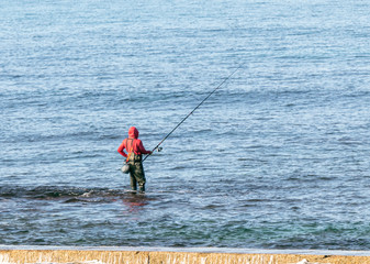 A fisherman  in waterproof clothing stands knee-deep in the water of the Mediterranean Sea and catches a fish on a fishing rod near the Roman aqueduct in Caesarea, in northern Israel.