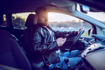 Young handsome caucasian bearded blond man in leather jacket driving his car and using smart phone.
