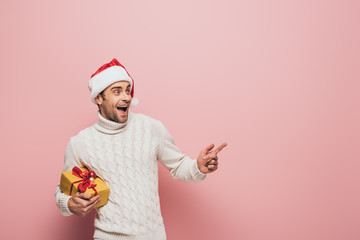 Fototapeta na wymiar handsome excited man in santa hat pointing and holding christmas present, isolated on pink