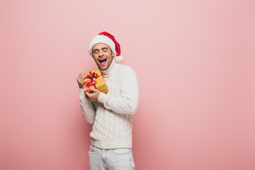 excited man in santa hat holding christmas gift, isolated on pink