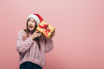 emotional woman in sweater and santa hat holding christmas gift, isolated on pink