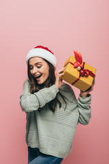 excited woman in sweater and santa hat holding christmas gift, isolated on pink