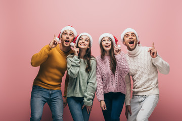 happy friends in sweaters and santa hats pointing up, isolated on pink