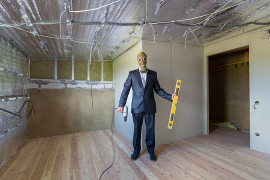 Strange man in businessman suit and gas protection mask inside a room under renovation works holding electric screwdriver and a level tools.