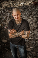 A young handsome bald brutal guy stands against the backdrop of the ruins of a building. Tattooed attractive man with arms fully covered by tattoos in a ruined building. Concept of crime, hooliganism.