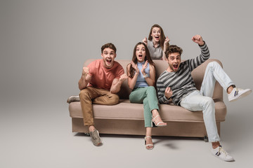 happy young friends cheering and sitting on sofa on grey