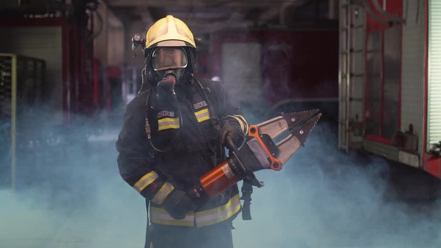 firefighter portrait wearing full equipment, oxygen mask, and power hydraulic cutting tool, smoke and fire trucks in the background. Name tag says fireman and model's last name.
