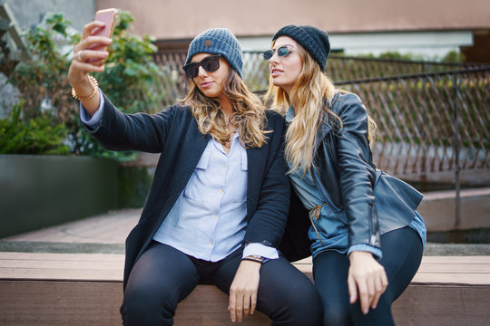 Two young happy girls sitting on a bench making selfies. Best friends having fun making self pictures with the smartphone wearing hats and sunglasses.