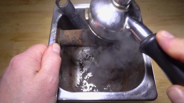 Slow motion overhead shot of a man&rsquo;s hands knocking steaming espresso coffee grounds from a commercial portafilter onto the padded bar of a stainless steel knock-off container / knock box.