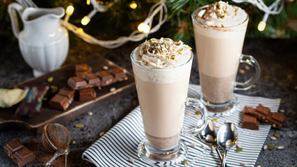 Christmas latte with whipped cream and pieces of chocolate with spoons on a napkin on a Christmas tree lighting background