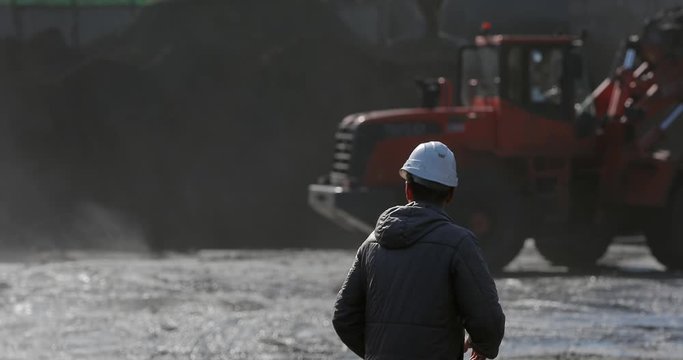 Marine coal terminal. A port worker in a white helmet stands with his back and watches the coal terminal work.