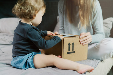 Fototapeta na wymiar Baby and woman sitting on bed and opening a mail box with products ordered on Internet. Online retailers offering wide range of goods delivered to your home address. Safe and quick shipping worldwide.